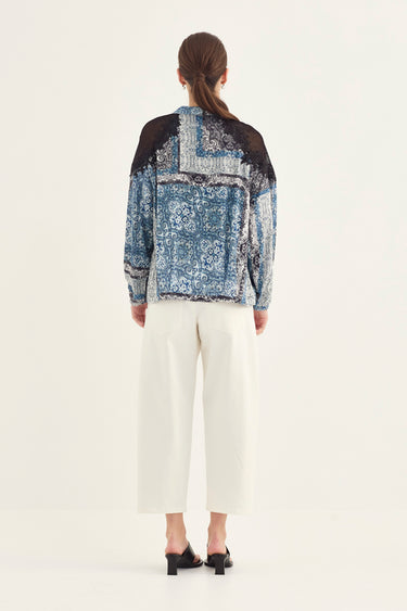A woman viewed from the back wearing a long-sleeve patchwork jacket with blue and white patterned fabric, black lace panels, and white cropped pants by Verge against a plain light background.