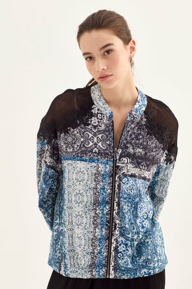 A woman wearing a patterned zip-up jacket by Verge with blue, white, and black paisley and damask prints, featuring sheer black mesh shoulder panels, photographed against a plain off-white studio background.