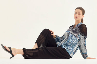 A woman sits on a white studio floor wearing a blue and white patterned blouse with black sheer sleeves, black wide-leg pants by Verge, and black high-heeled sandals.