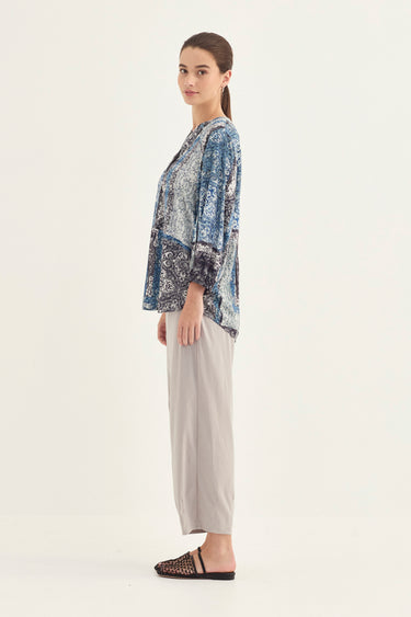 A woman stands in profile wearing a loose blue and white patterned top with light beige wide-leg cropped pants by Verge and dark woven slide sandals, photographed against a plain light background.