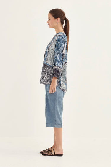A woman shown in profile wearing a blue and white patterned long-sleeve top, light blue cropped jeans by Verge, and black flat mules against a plain off-white studio background.
