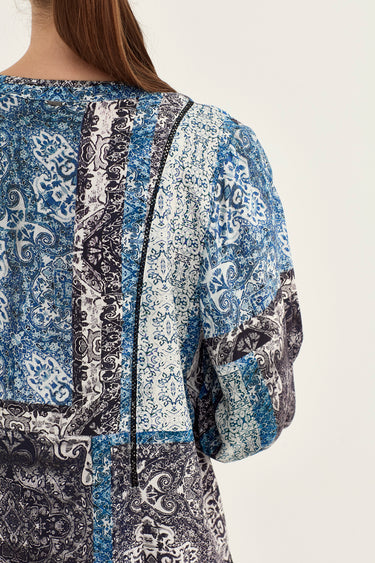 Close-up back view of a woman wearing a blue, white, and black paisley patchwork blouse by Verge against a light beige background.